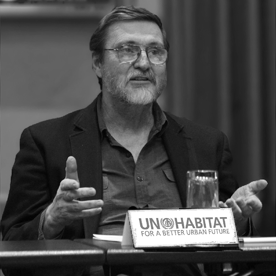 man with glasses at a desk, looking like he is in mid-speech. A sign on desk says "UN Habitat for a better urban future"