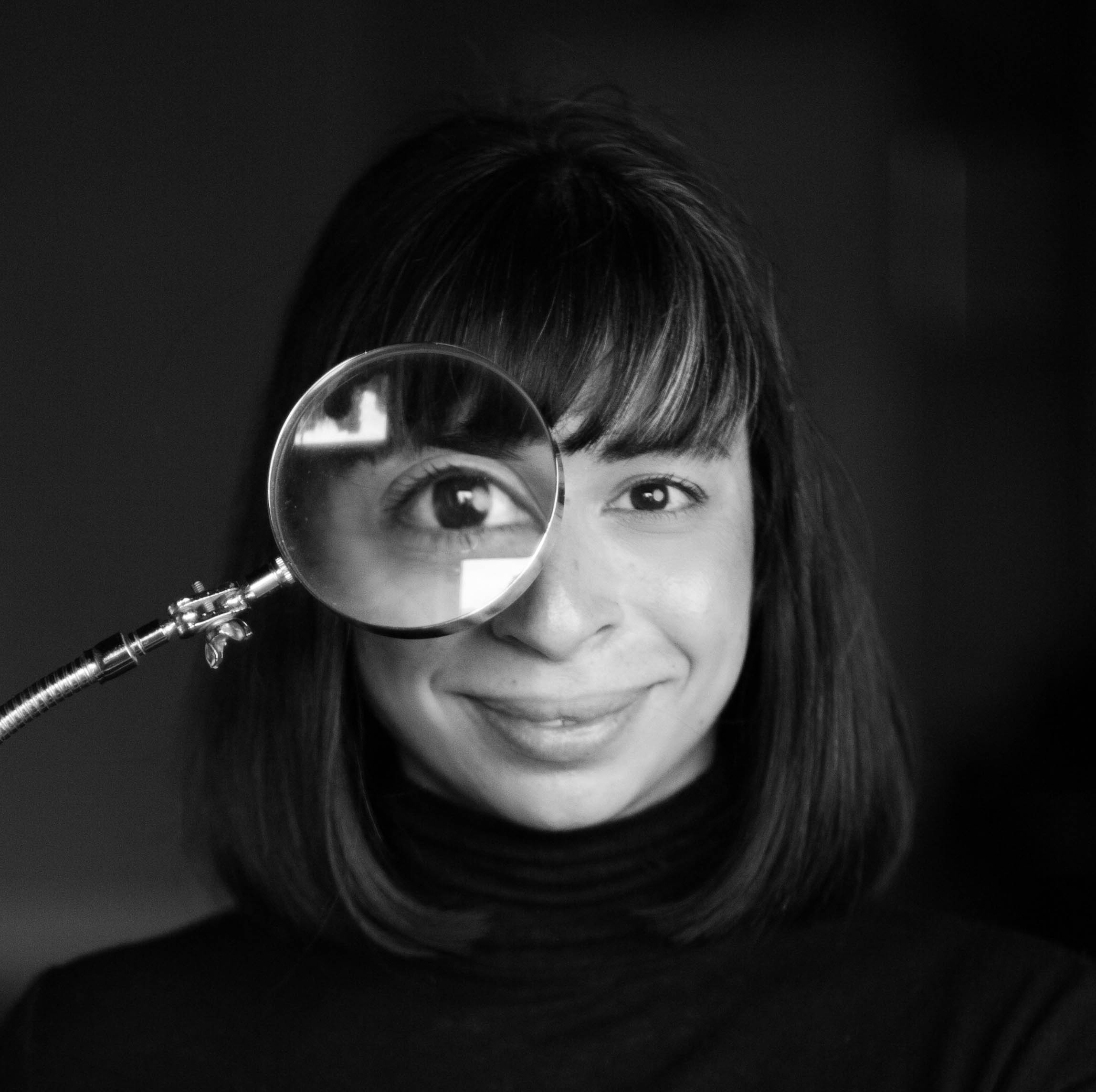 headshot of woman with dark bangs and shoulder length hair, holding a magnifying glass over one eye