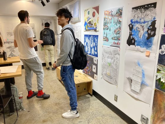 Students stand in a room with white walls covered in posters with student drawings