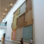 People look at a large piece of art on a wall. This piece looks like a giant wooden quilt.
