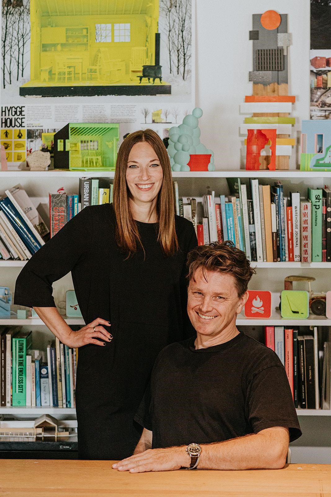 Woman standing beside a seated man, both smiling at the camera. A bookshelf and books in the background