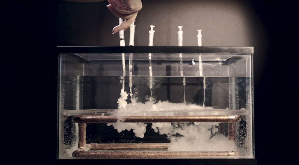 five syringes containing an undisclosed white liquid are being disposed into a tank of clear water with a brown bar of copper at the bottom of the tank