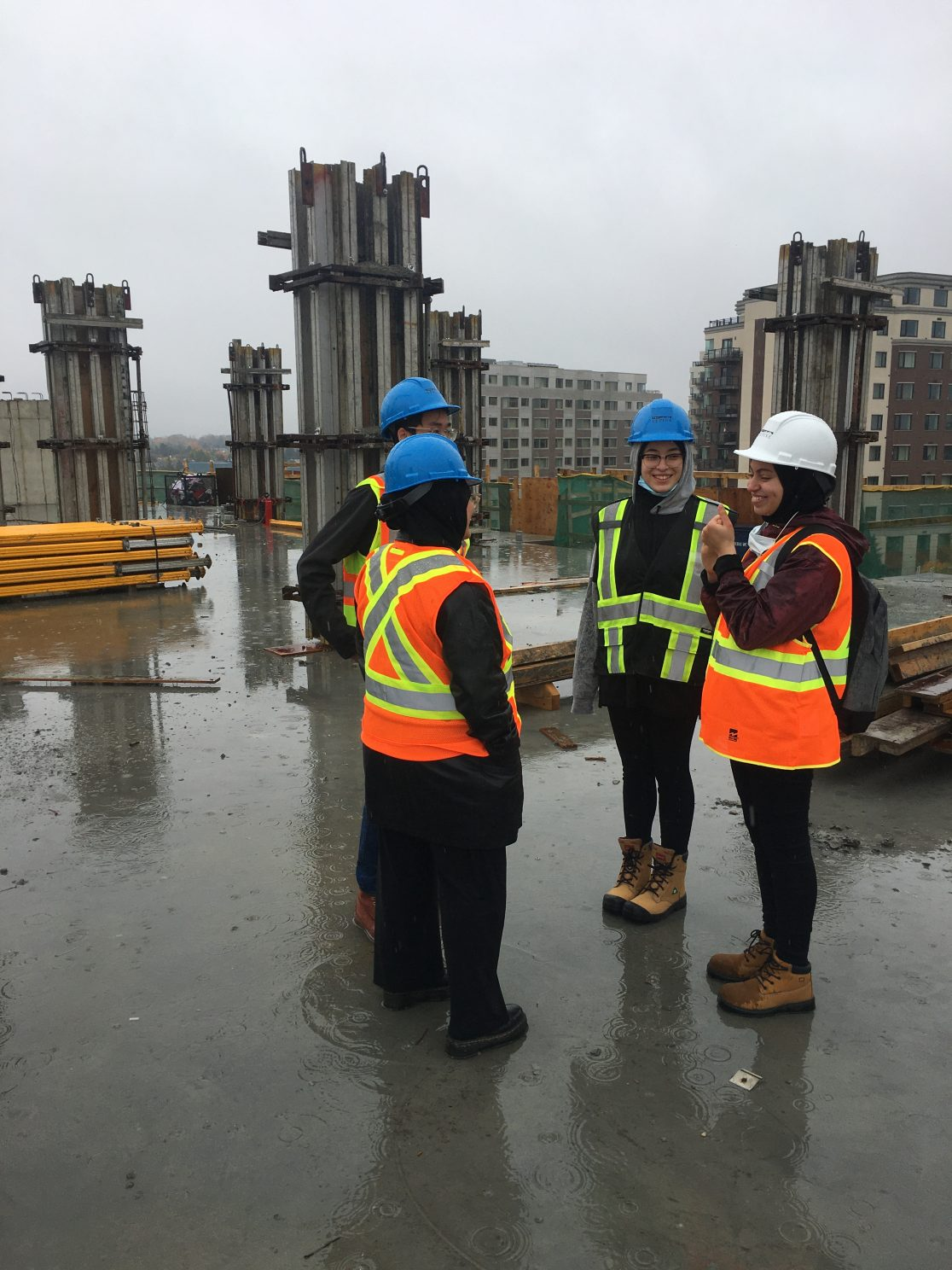 4 students talking in a circle wearing construction vests and hats on a construction site.