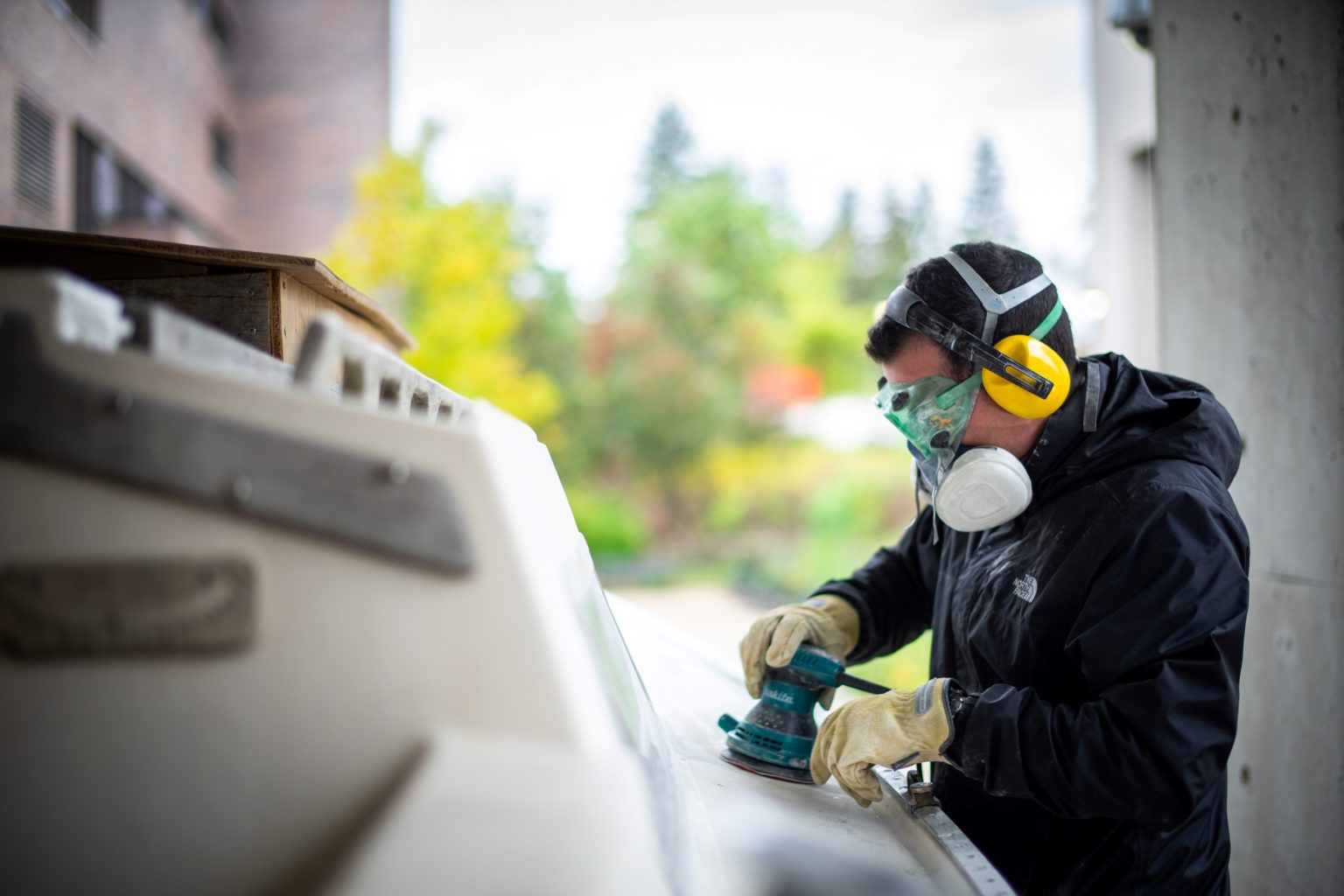 A man waring a respirator mask and noise cancelling earmuffs sands down a sailboat.