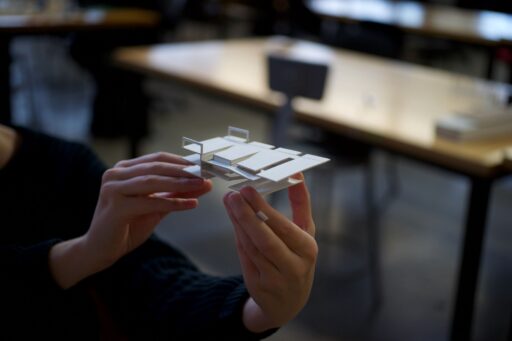 Hands holding a small architectural model, with tables and a classroom setting blurred in the background.