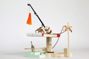 A model constructed from wood, featuring animal figurines and a black rod holding an orange tassel.