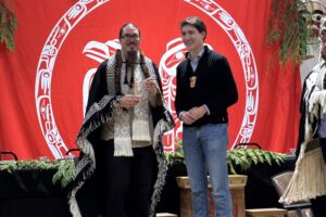 A Haida representative holds a small copper pine tree while standing beside Prime Minister Justin Trudeau in front of a red Haida Nation banner