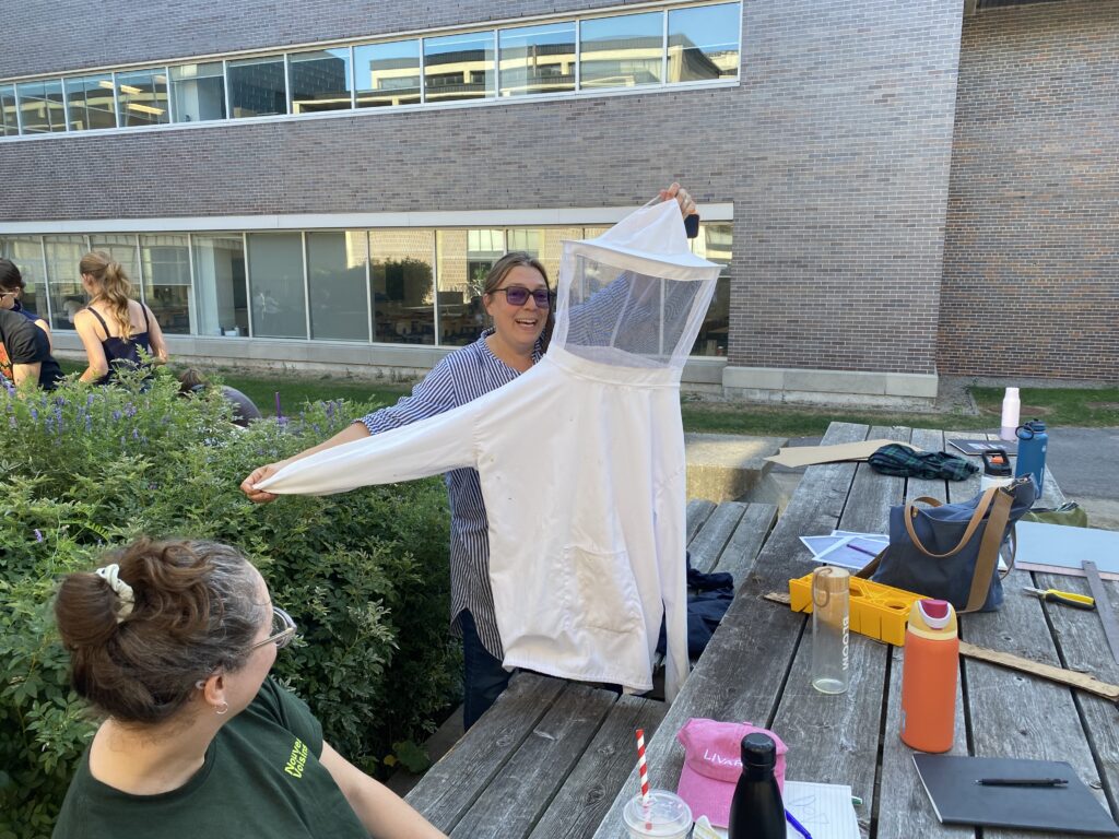 Debanné holding up a white beekeeping suit beside a picnic table covered with tools and drinks.