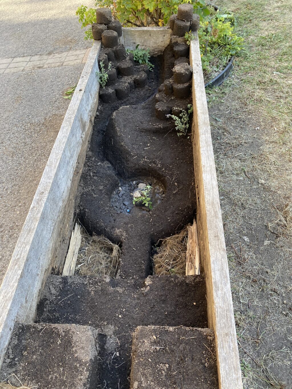 Wooden planter with carved earth path and stacked cylindrical soil blocks along the sides.