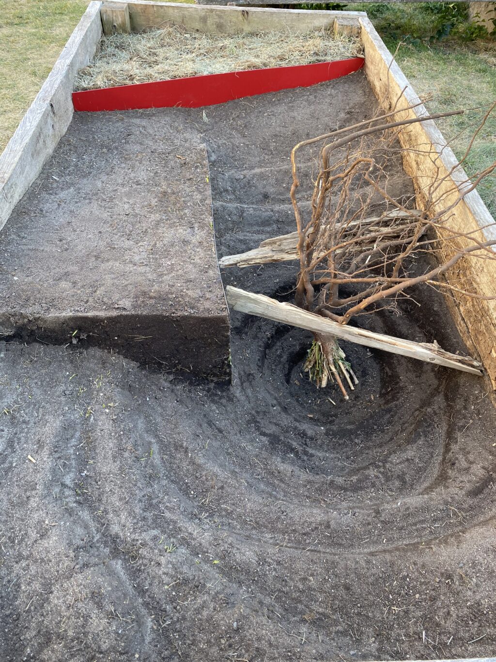 Curved earthwork planter with swirling soil pattern, red divider, and dry branches anchored in the center.