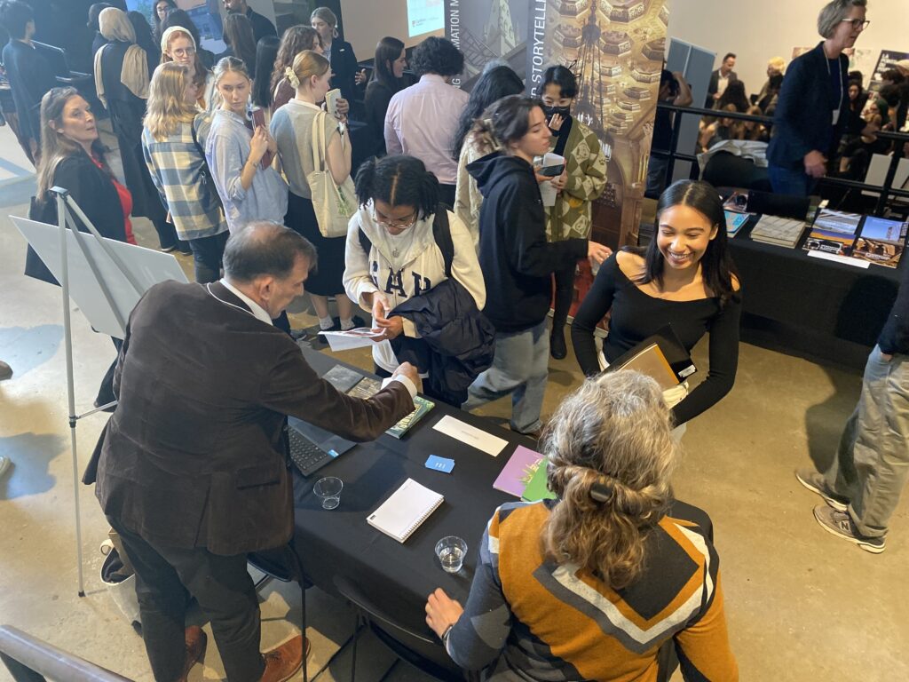 Students speaking with two seated employers at a career fair table, surrounded by others in conversation.