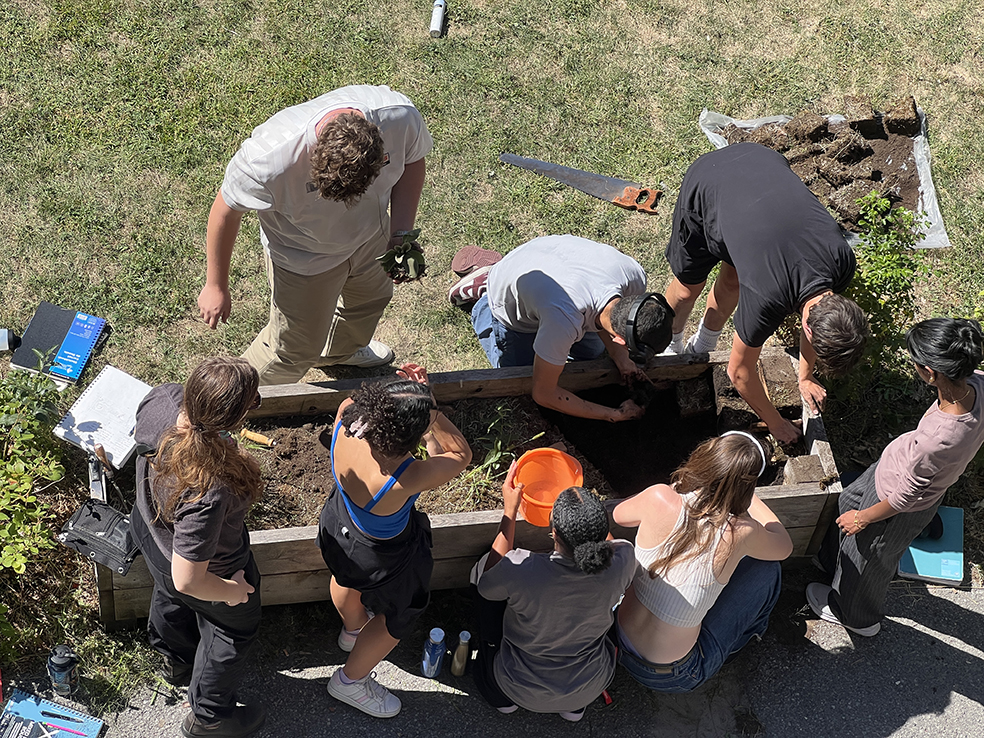 Overhead view of a group of students kneeling and standing around a wooden planter, digging and shaping soil.