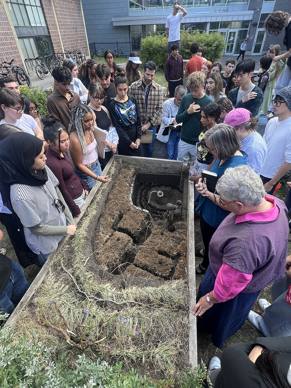 Students and instructors gather around a sculpted earthwork inside a wooden planter filled with carved soil and grasses.