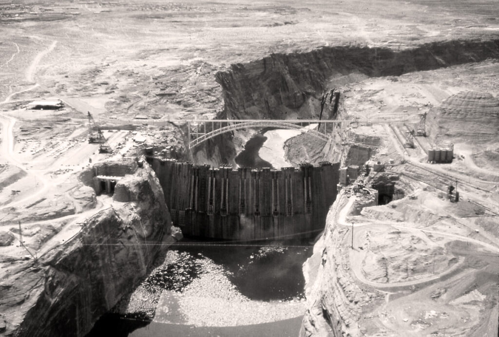 Aerial view of Glen Canyon Dam spanning the Colorado River, with Glen Canyon Bridge and desert landscape beyond.
