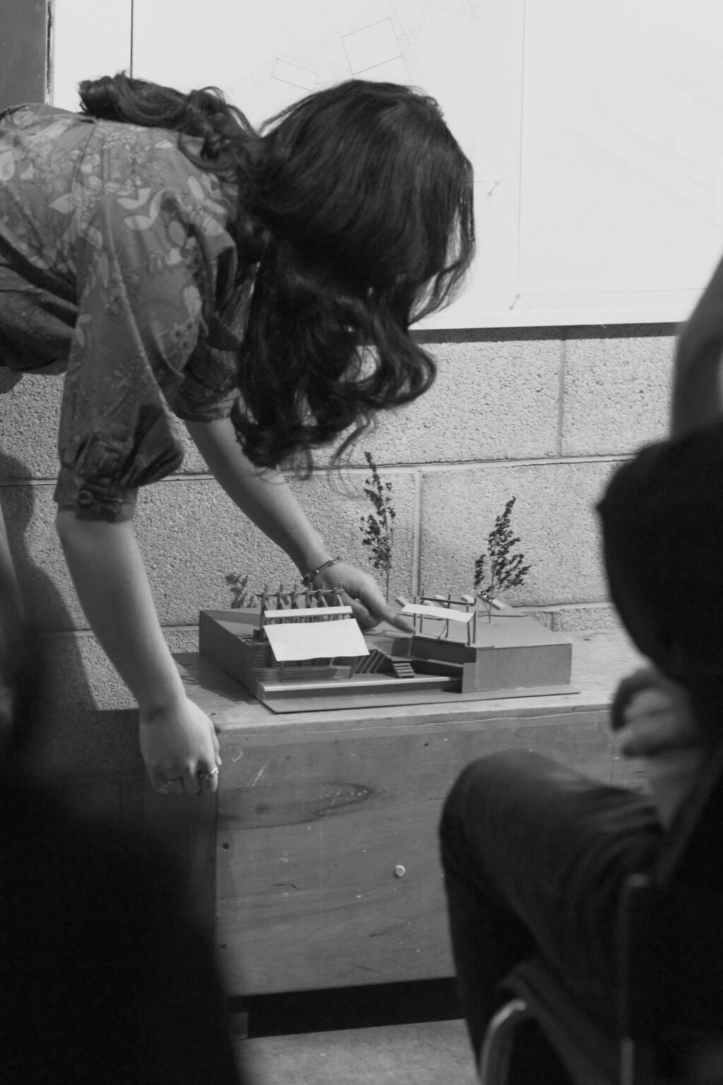 Student leaning forward to adjust an architectural model placed on a wooden table during a presentation.