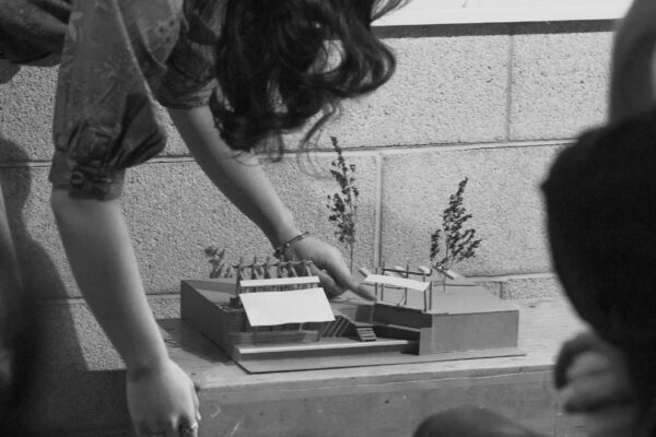 Student leaning forward to adjust an architectural model placed on a wooden table during a presentation.