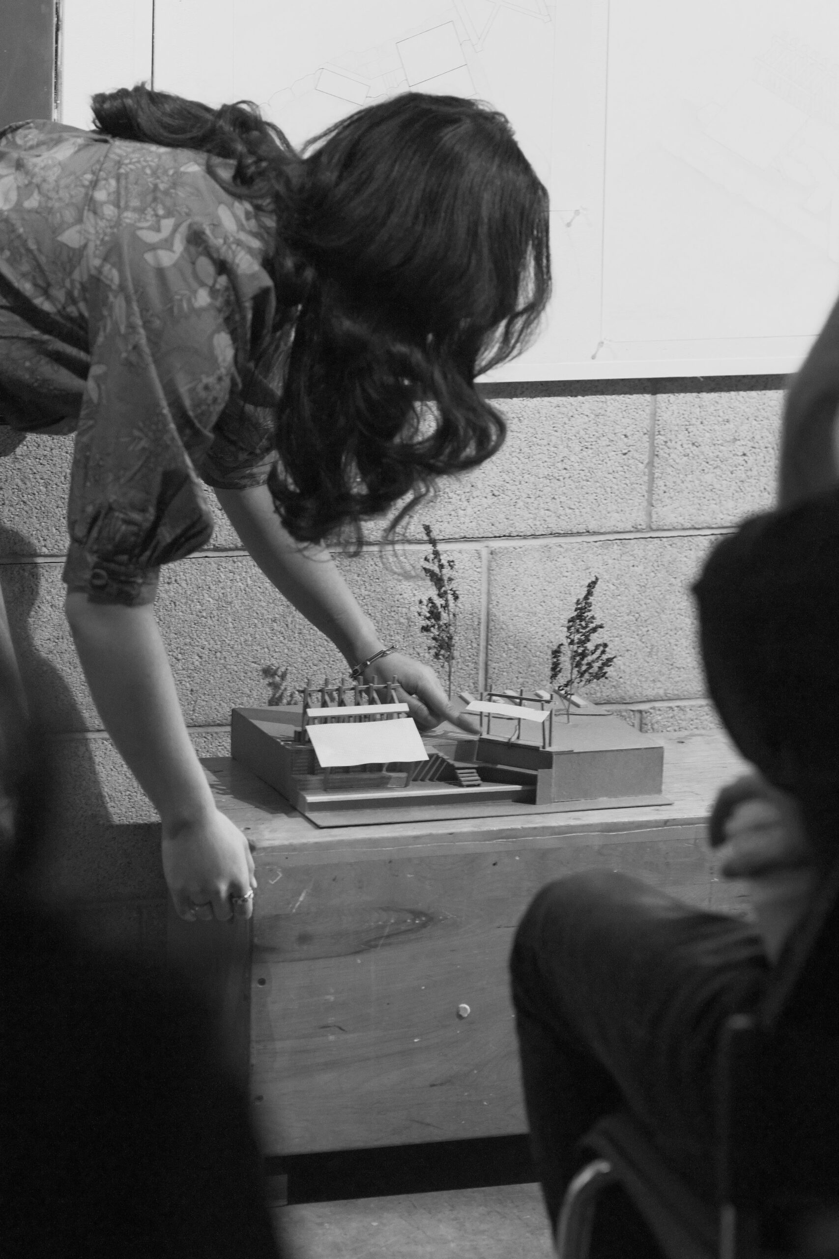 Student leaning forward to adjust an architectural model placed on a wooden table during a presentation.