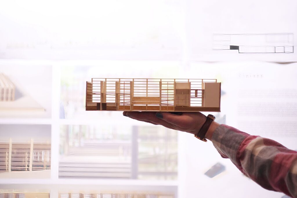 Hand holding a small wooden architectural model in front of a wall display with drawings
