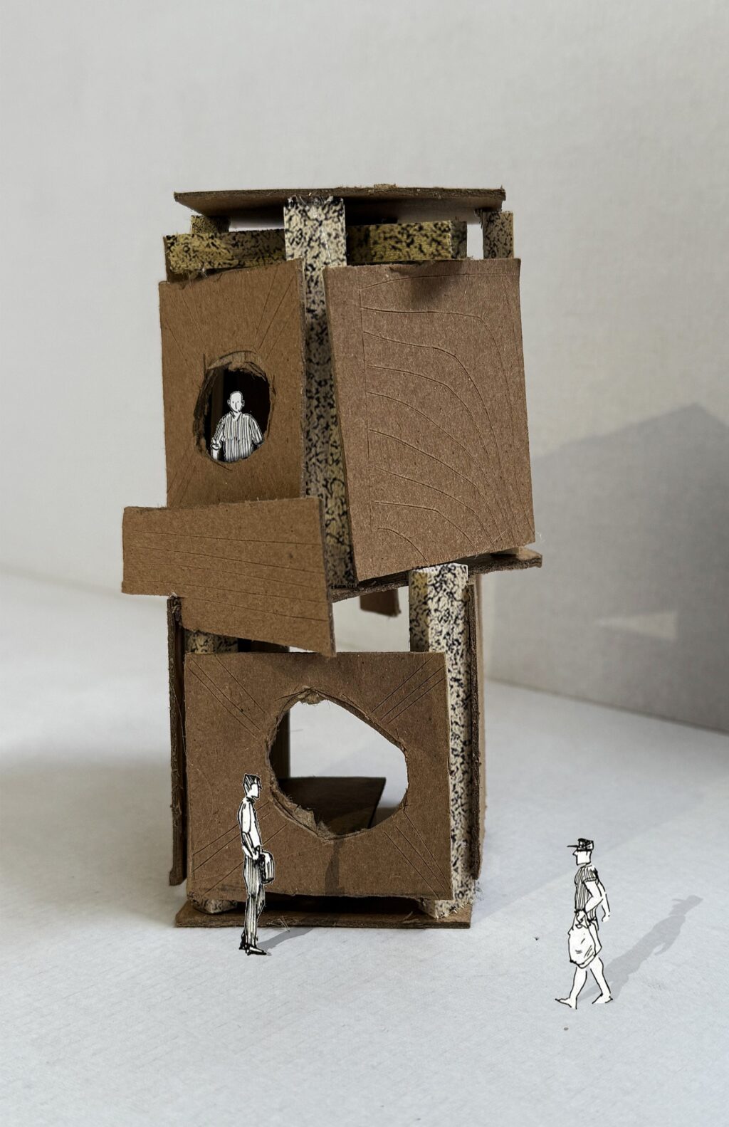Cardboard architectural model with cut-out windows and miniature paper figures standing around it on a white background.