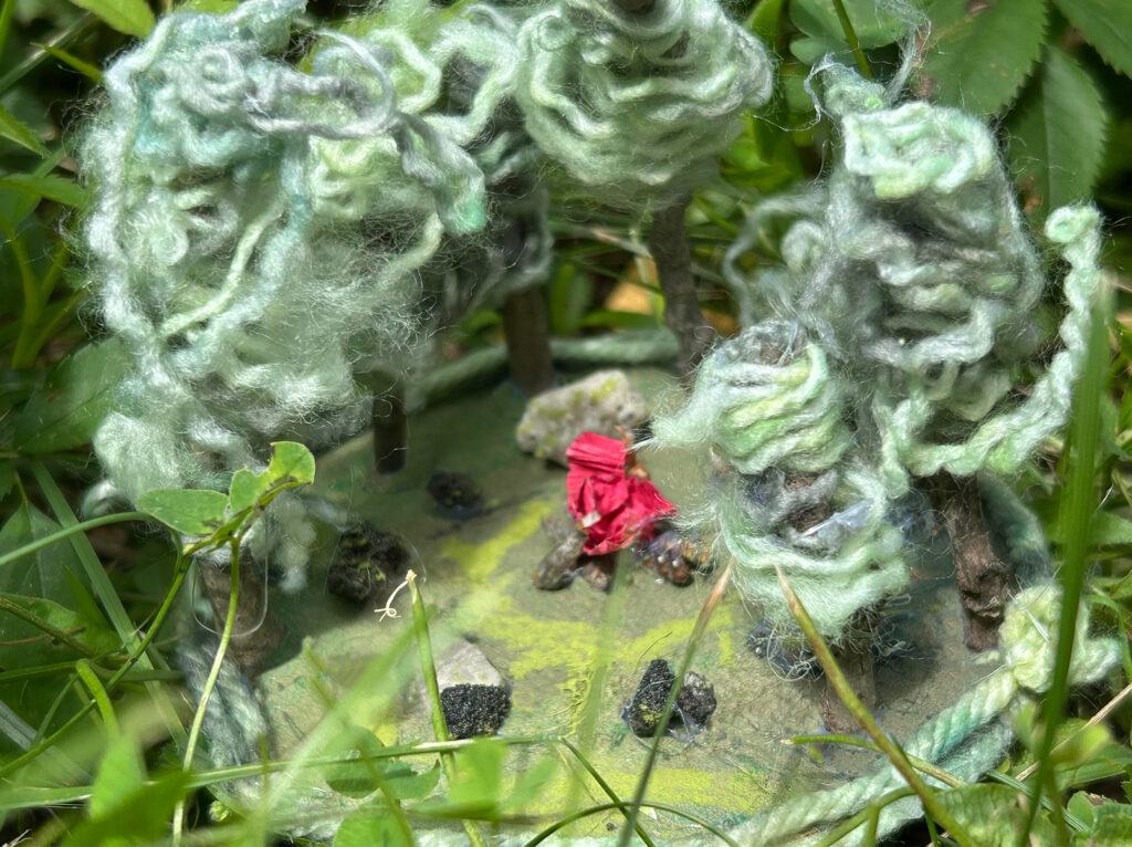 Miniature forest scene with yarn trees and a red paper figure, surrounded by grass and natural ground.
