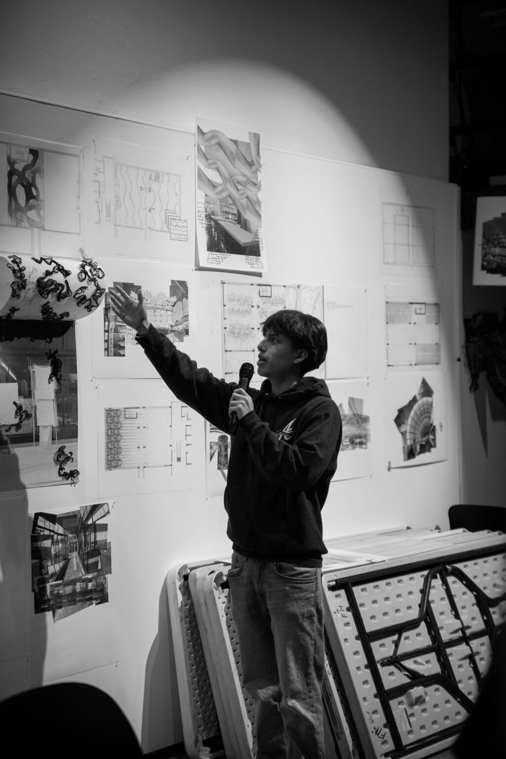 Student holding a microphone and pointing toward architectural drawings pinned on a wall during a presentation.