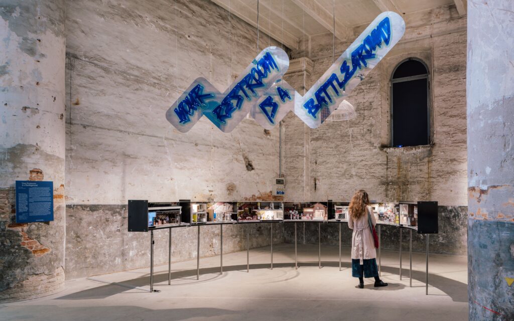 Person viewing architectural models beneath suspended blue signage reading “YOUR RESTROOM IS A BATTLEGROUND.”