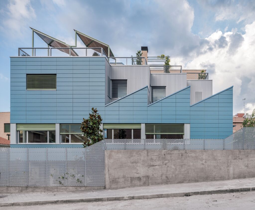 Blue multi-storey building with sawtooth roof profile, green window frames, and rooftop terraces against a cloudy sky.