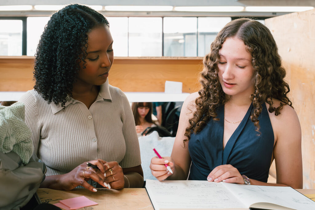 Two students sitting at a desk, one showing sketches in a notebook to the other.