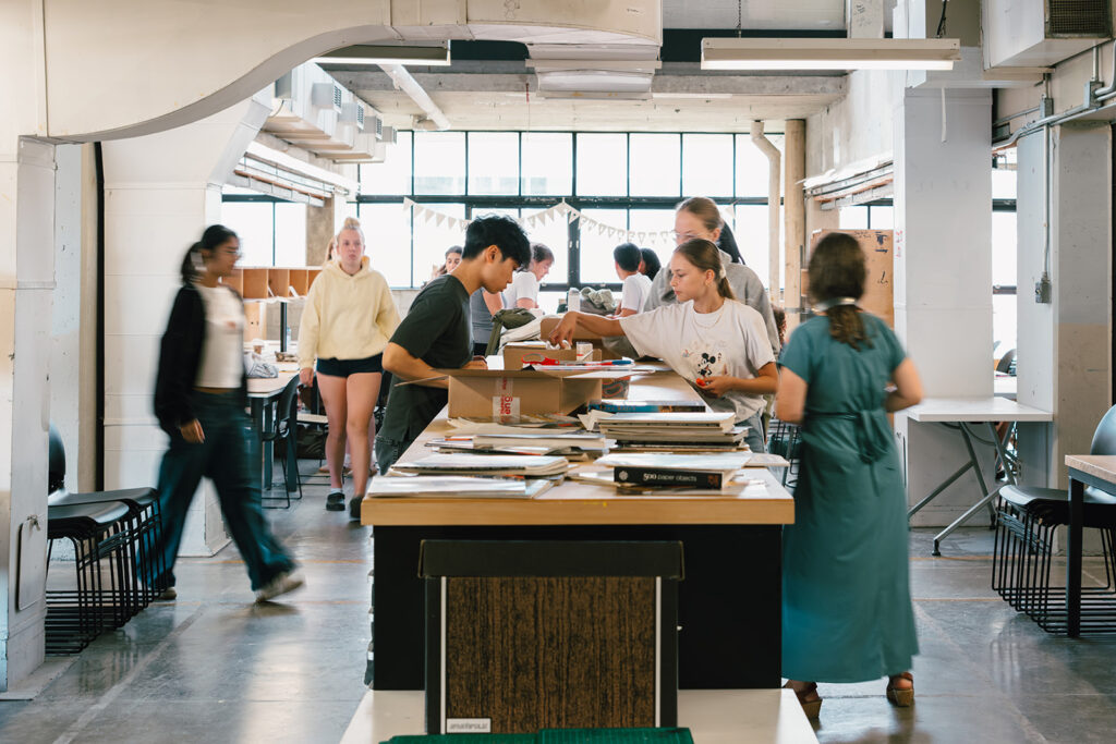 Students gather around a large table in a studio, sorting through cardboard boxes and supplies.