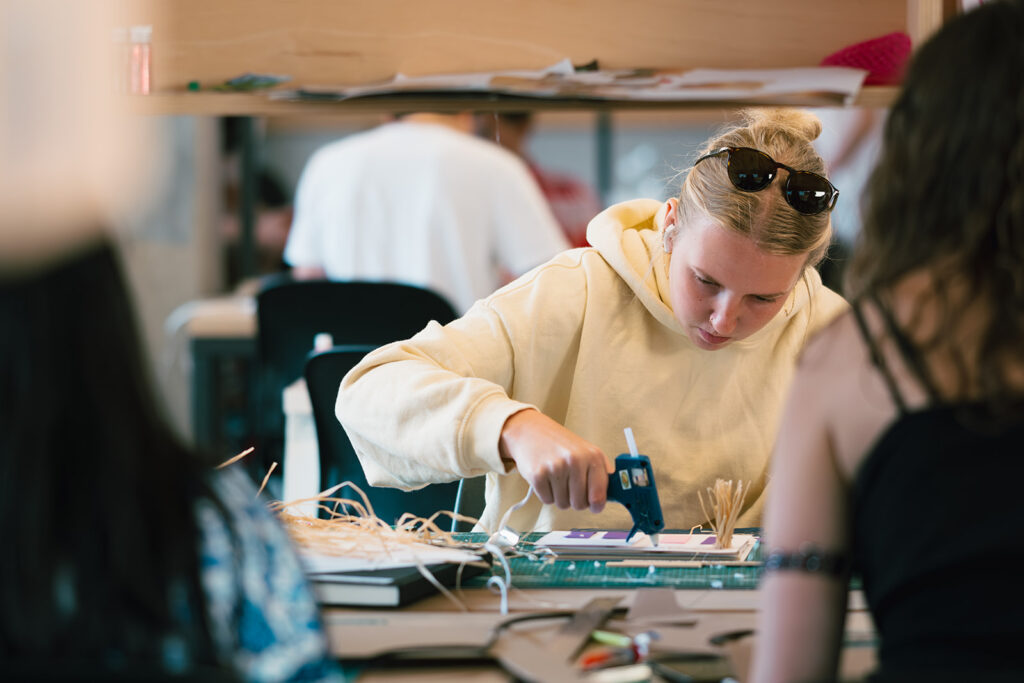 Student using a glue gun to build a model at a desk covered with materials.