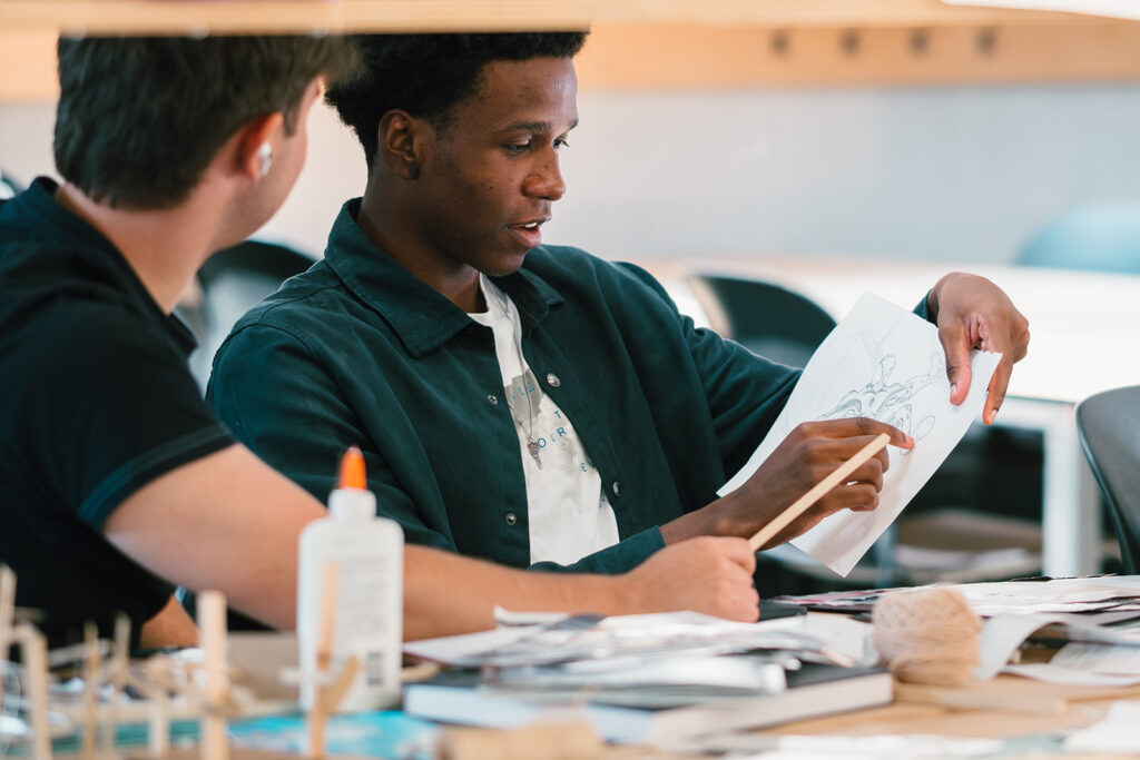 Two students sitting at a studio desk, discussing a hand-drawn diagram with modeling supplies around.