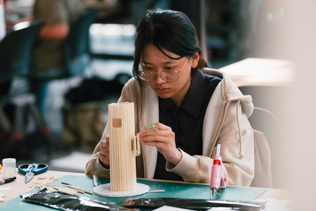 Student working with wood sticks to construct a vertical architectural model on a green cutting mat.