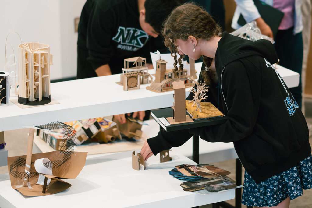 Student places small architectural models made of cardboard onto a white table for display.