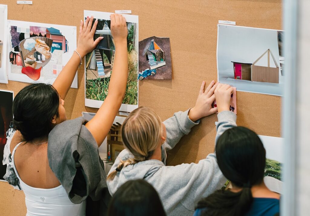Students pin printed architectural collages and model images onto a display board.