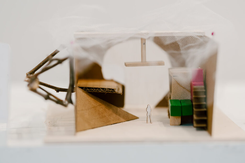 Close-up of a cardboard architectural model with twig structure, mesh roof, and small interior furniture pieces.