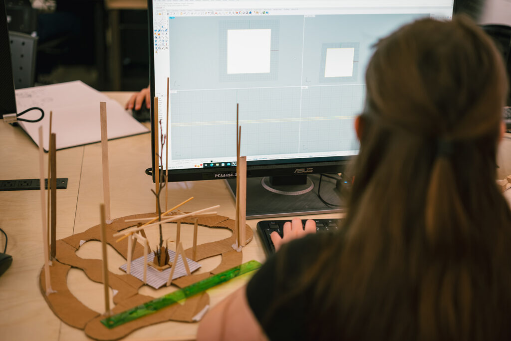 Student working in Rhino on a desktop computer next to a cardboard and wood architectural model.
