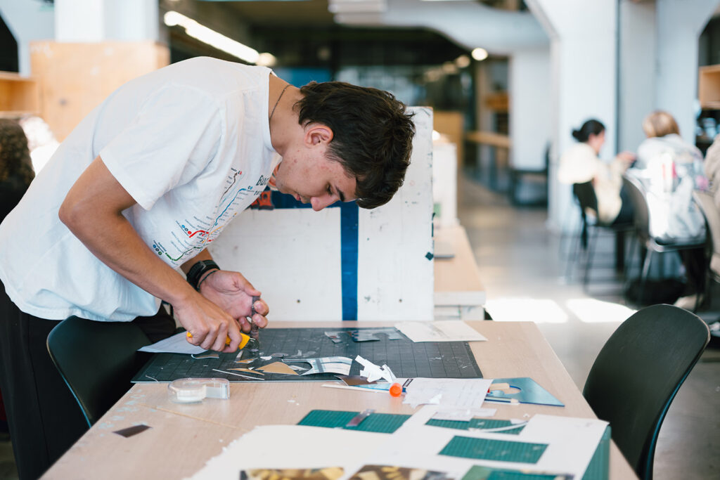 Student cuts paper pieces with a craft knife at a studio table.