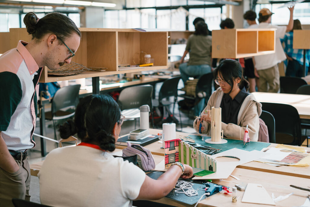 Student explains her architectural model to another person at a shared studio table.