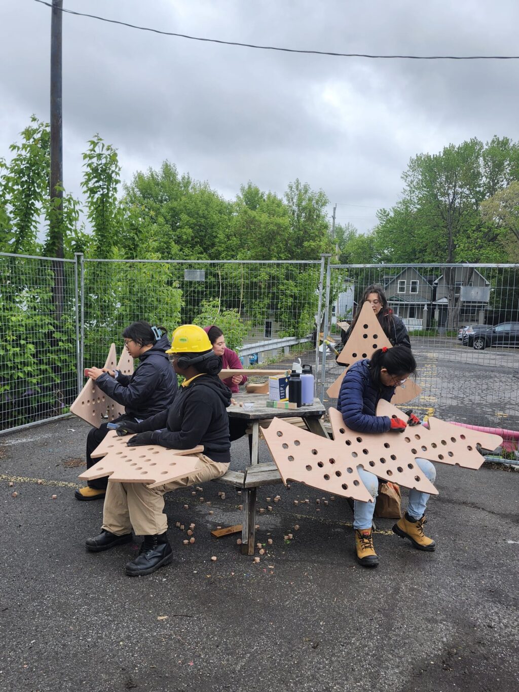 Students sanding and assembling perforated wooden tree panels outdoors at the Vanier HUB build site.