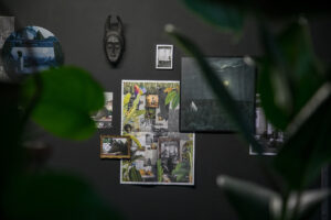 Installation view of a black wall featuring a collage of photographs, a dark mask with horns, and plant leaves in the foreground.