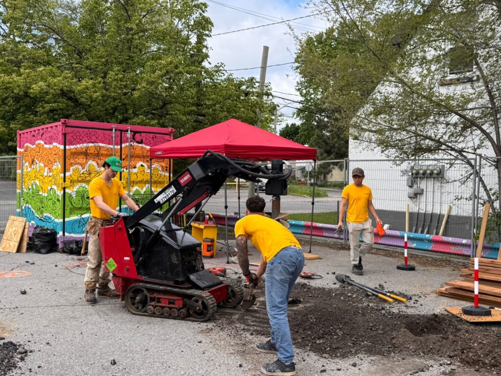 Students using a compact loader on site near a colorful storage container and red tent.