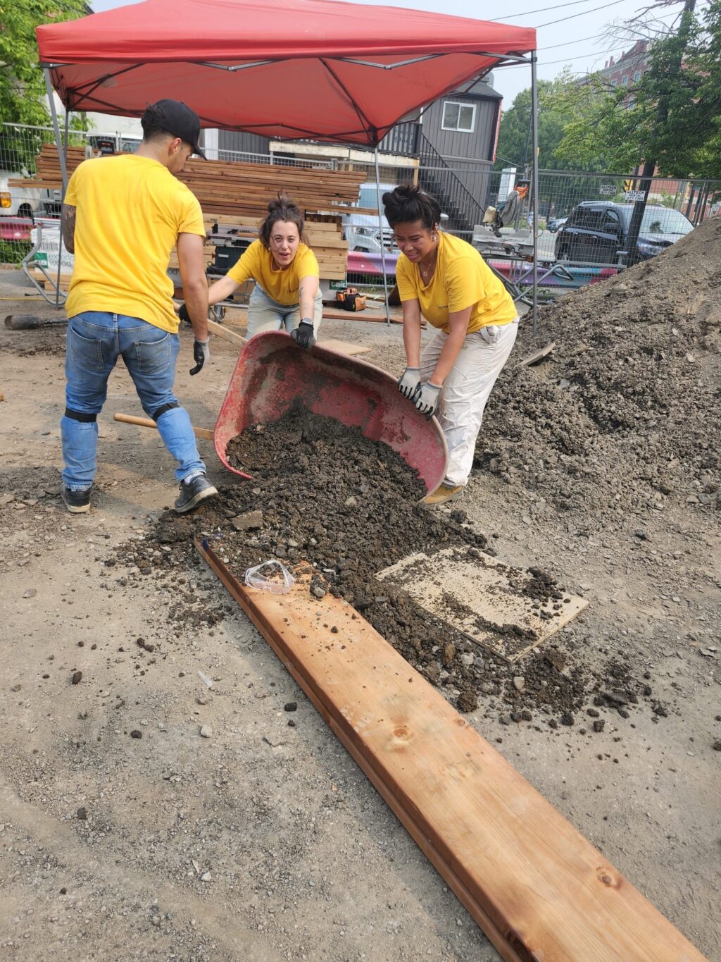 Students emptying a wheelbarrow of soil at the construction site under a red canopy.
