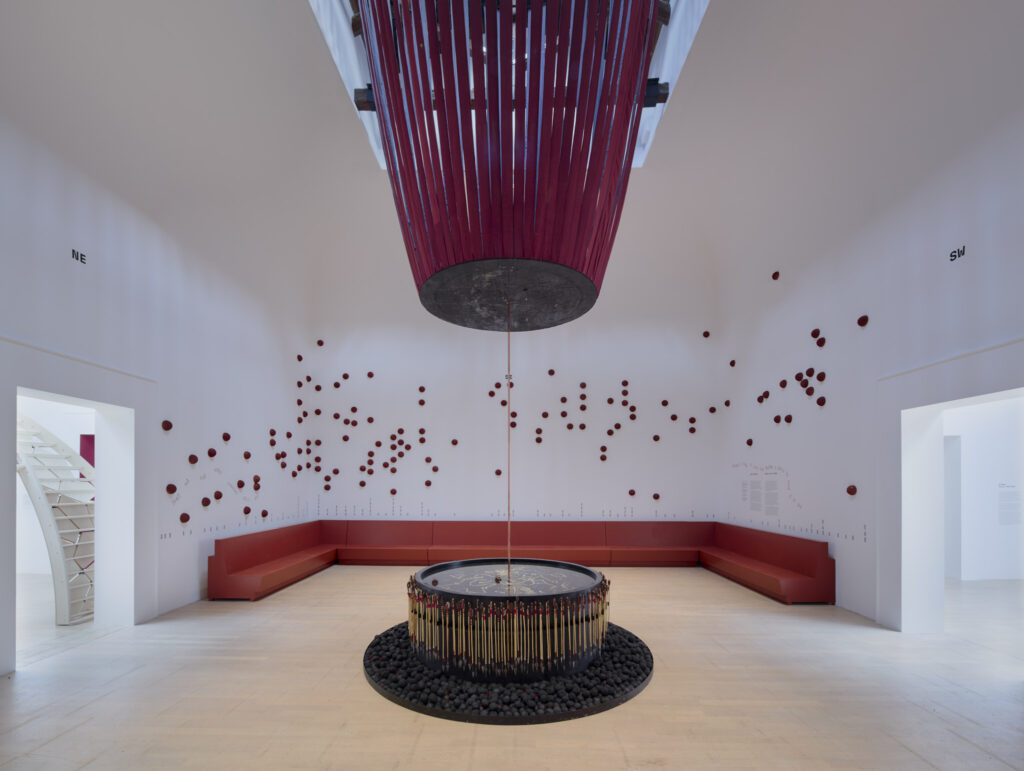 Interior installation showing a suspended red form above a circular map-like pool with wall markers.