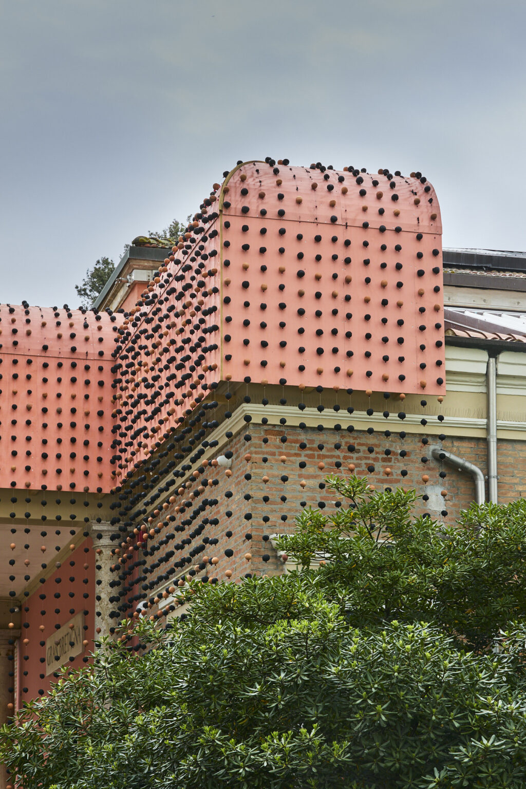 Close-up of a pavilion facade clad in pink panels and brick, studded with rows of black and terracotta circular knobs.