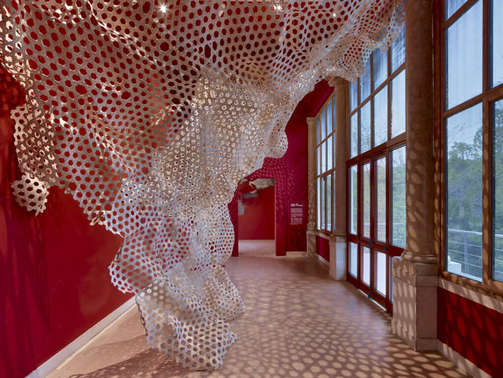 Perforated white metal installation hangs along a red-walled corridor, casting patterned light and shadow on the floor.
