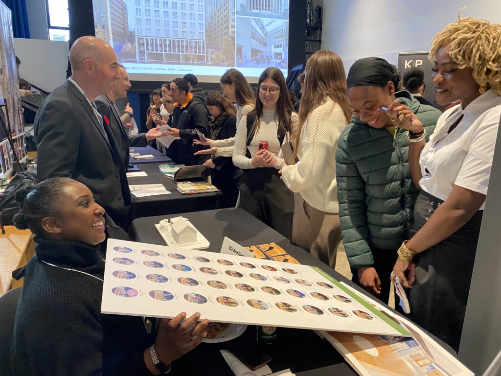 Camille Lewis holding a poster with circular portraits, seated at a recruitment table with students gathered around.