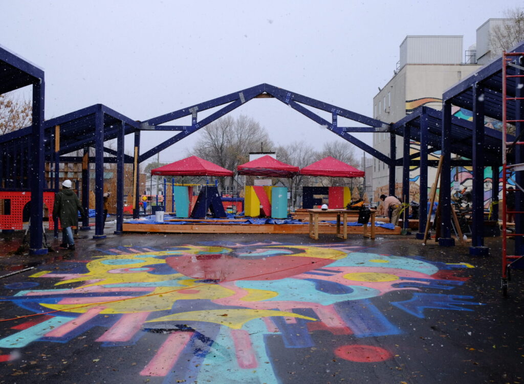 Snow falling over the colorful ground mural and blue open-frame structures during construction.