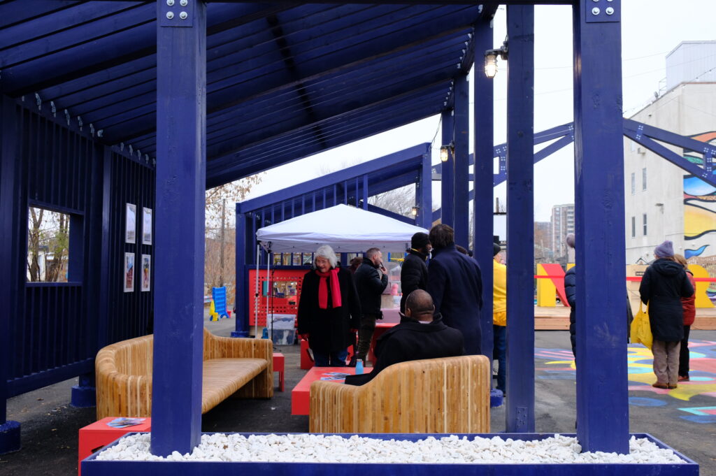 People gathering under the blue open-frame structure at Vanier Hub, with seating and displays nearby.
