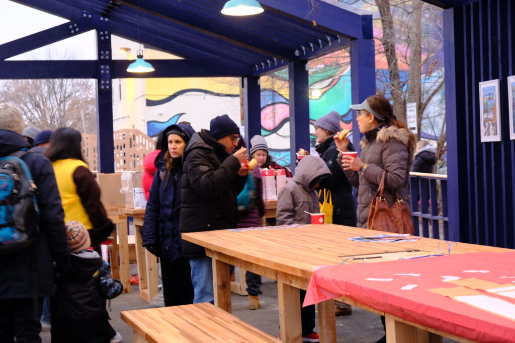 Visitors eating and gathering under the blue-framed structure with long wooden tables at the HUB opening event.