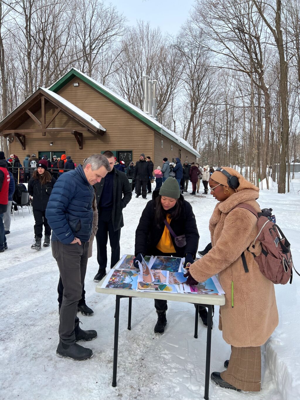 People standing around a table outdoors in the snow, looking through printed photos and discussing them.